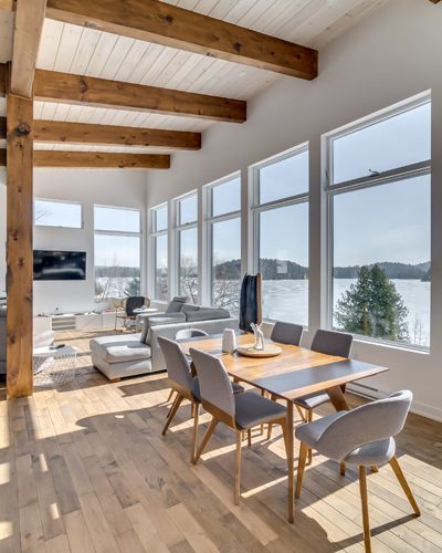 Salle à manger d'une maison Maisons Allaire : poutres de bois apparentes, plancher de bois franc et mur de fenêtres donnant sur un lac gelé
