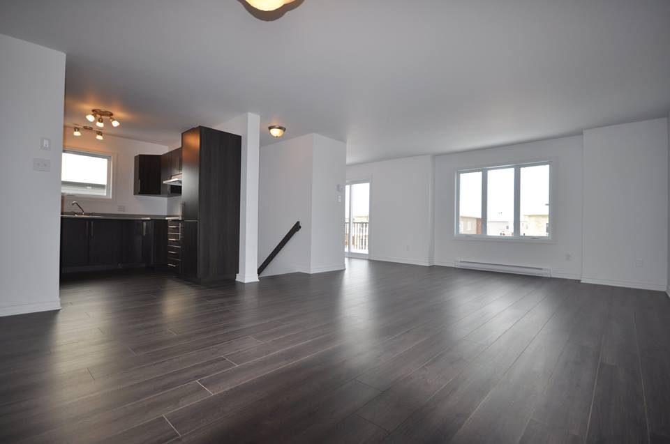 Intérieur d'un logement neuf : aire ouverte, cuisine sombre, plancher de bois foncé, deux fenêtres